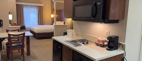 A hotel room with a kitchenette, including a microwave, stove, and coffee maker. There is a bed, a desk, and a chair.