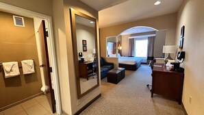 A hotel room with a bed, a desk, a chair, a sofa, a mirror, and a bathroom with a towel rack.