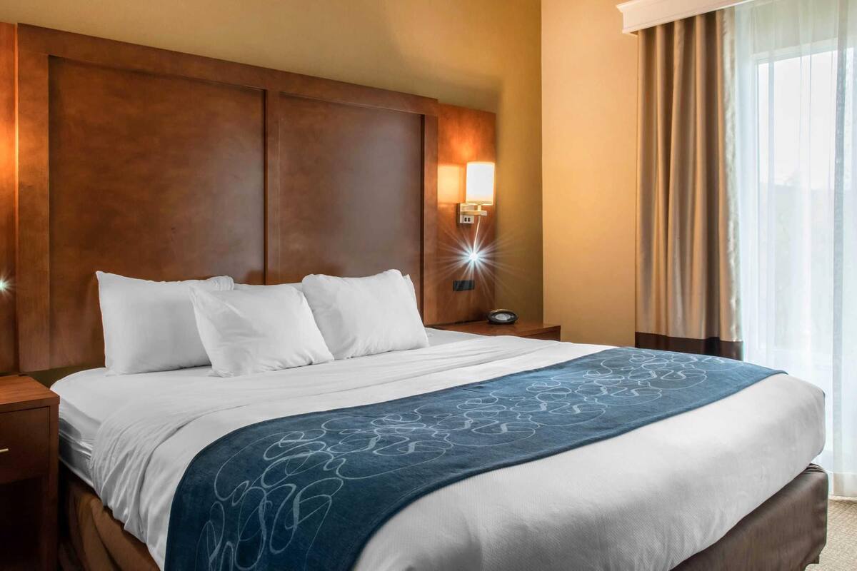 A hotel room with a large bed, a nightstand, a wall-mounted lamp, and a window with curtains.