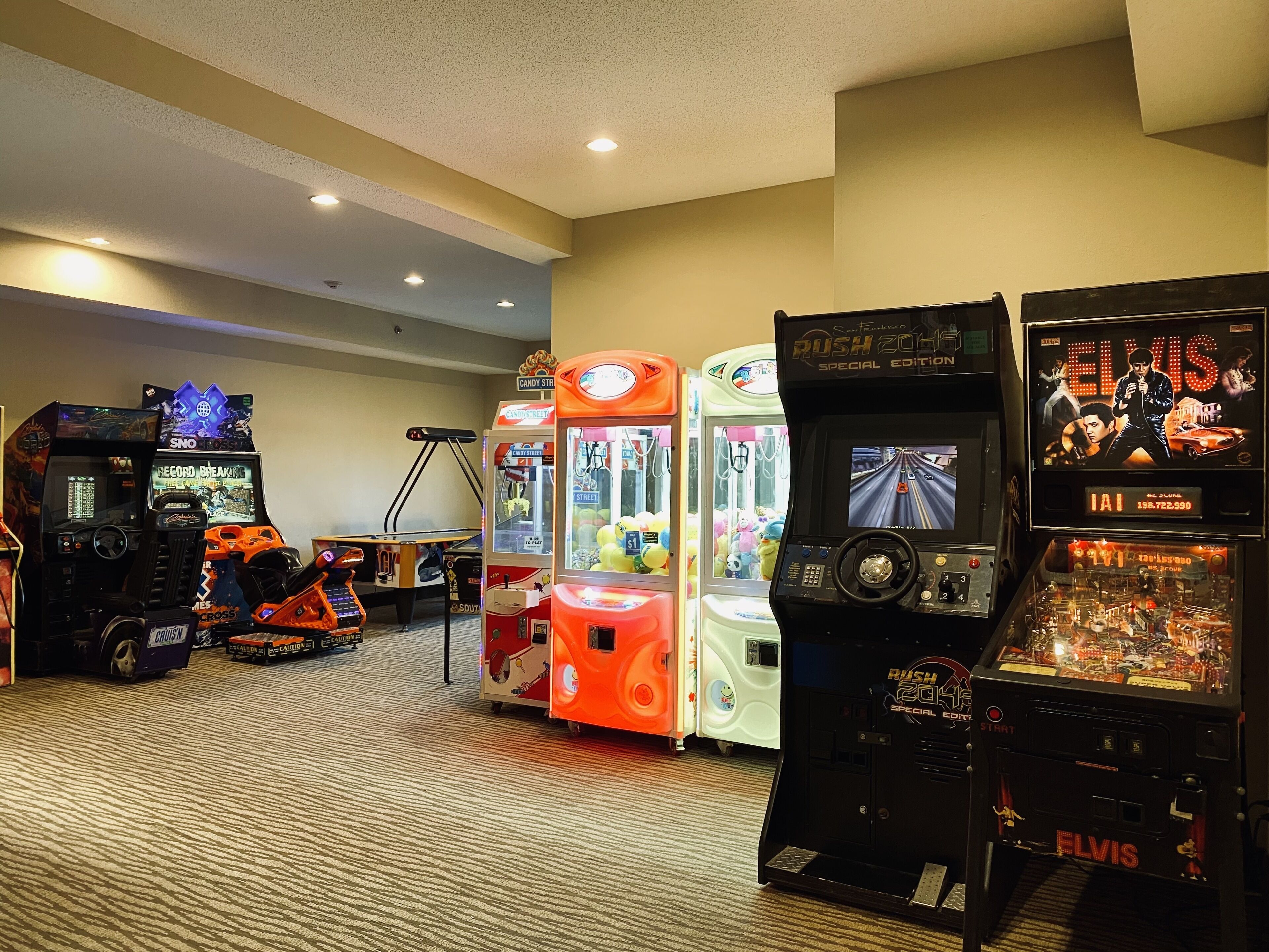 En rad arkadspel, inklusive ”Sno Cross”, ”Candy Street”, ”Rush 2048 Special Edition” och ”Elvis”, står uppställda i ett rum med heltäckningsmatta och infällda taklampor.