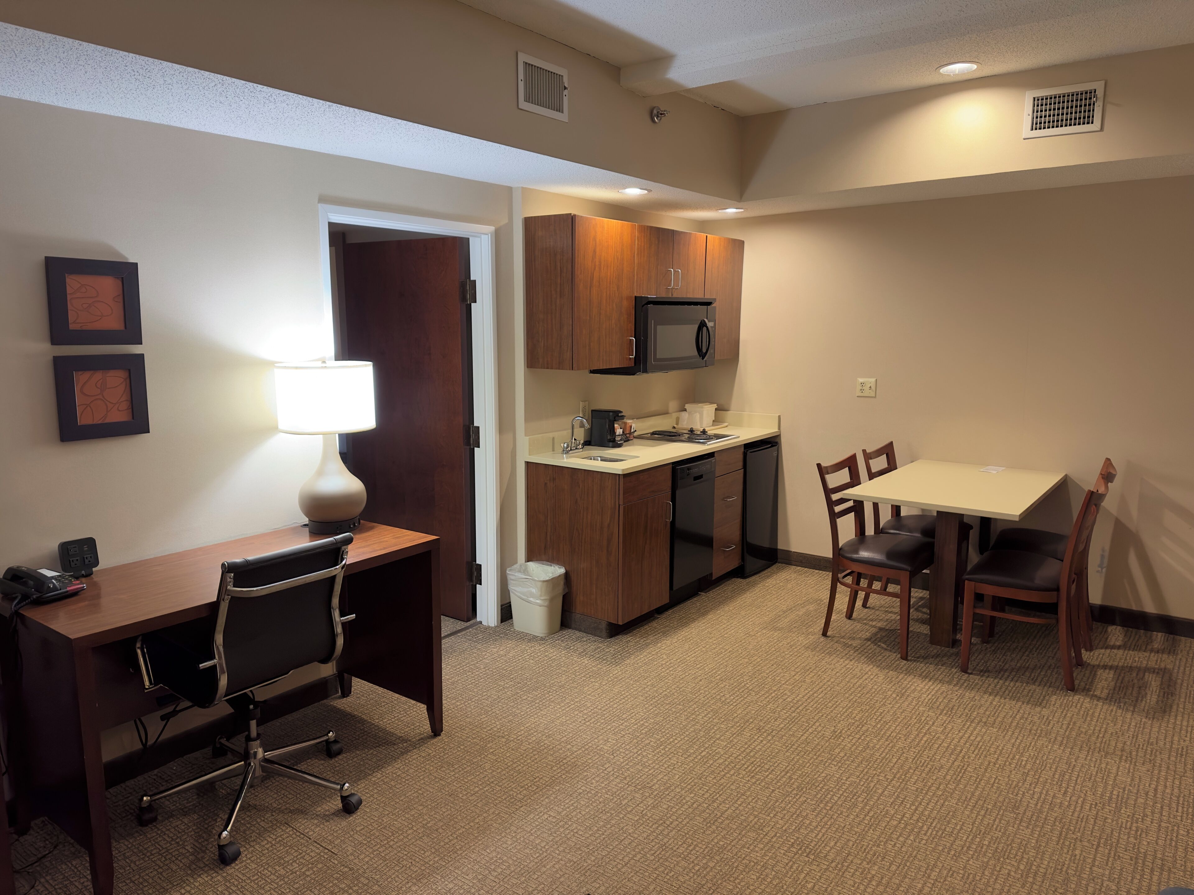 A compact hotel room with a kitchenette, a dining area, and a desk with a chair.