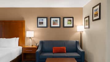 Quarto de hotel com cama, um sofá azul, mesa de centro de madeira, mesa de cabeceira com abajur e quadros nas paredes.