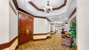 Un couloir d’hôtel avec un tapis à motifs, des portes en bois et un coin salon agrémenté d’une plante.