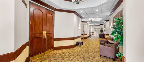 A hotel corridor with patterned carpet, wooden doors, and a seating area with a plant.