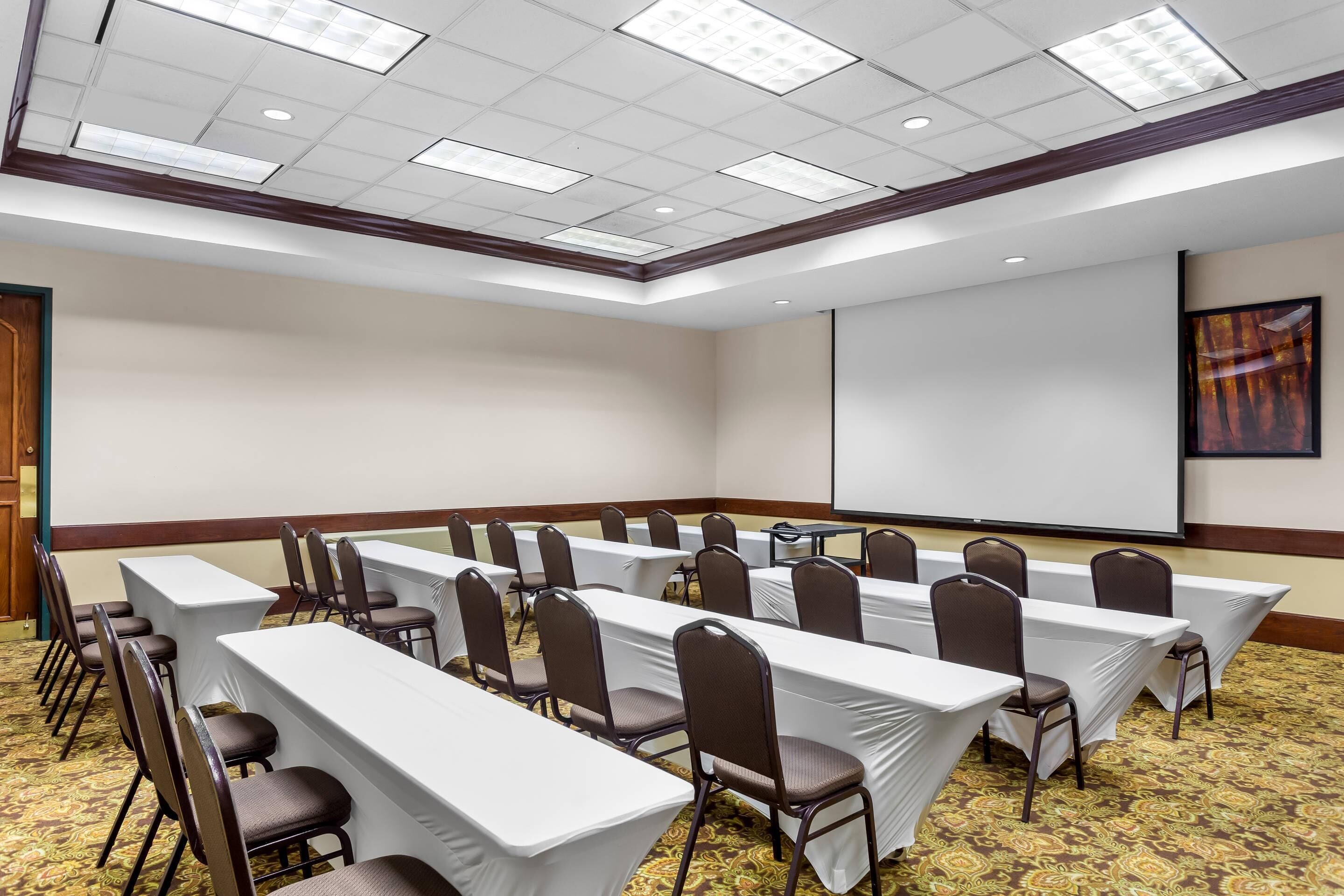 A conference room with long tables, chairs, a projector screen, and a carpeted floor.