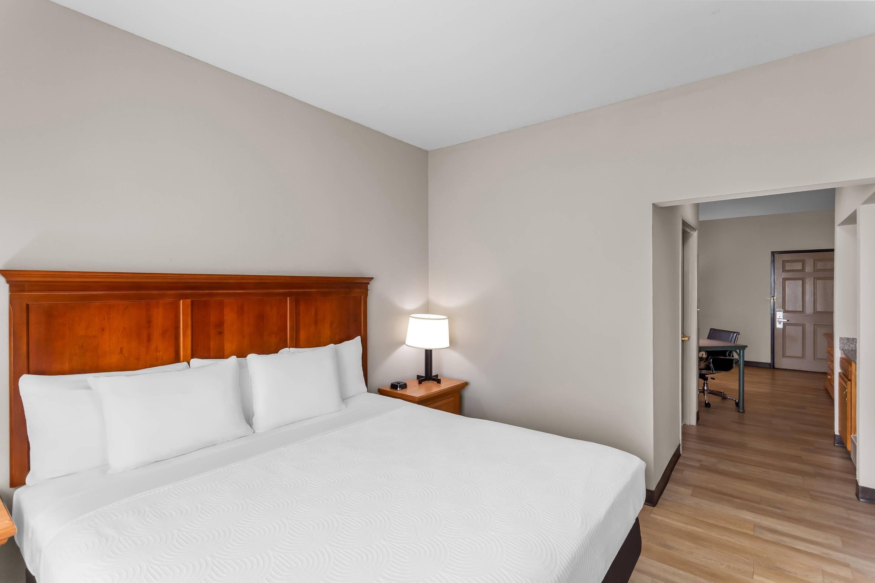 A hotel room with a large bed, a wooden headboard, a nightstand with a lamp, and a view of the hallway and another room.