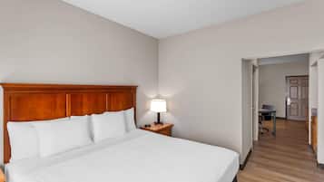 A hotel room with a large bed, a wooden headboard, a nightstand with a lamp, and a view of the hallway and another room.