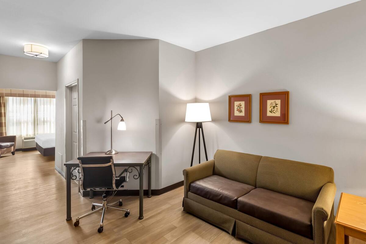 A hotel room with a wooden floor, a brown leather sofa, a desk with a chair, a standing lamp, and two framed pictures on the wall.