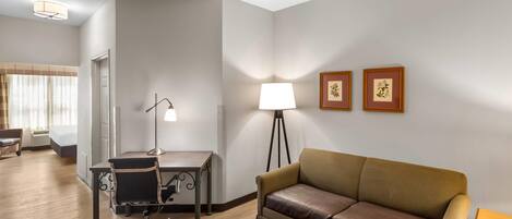 A hotel room with a wooden floor, a brown leather sofa, a desk with a chair, a standing lamp, and two framed pictures on the wall.