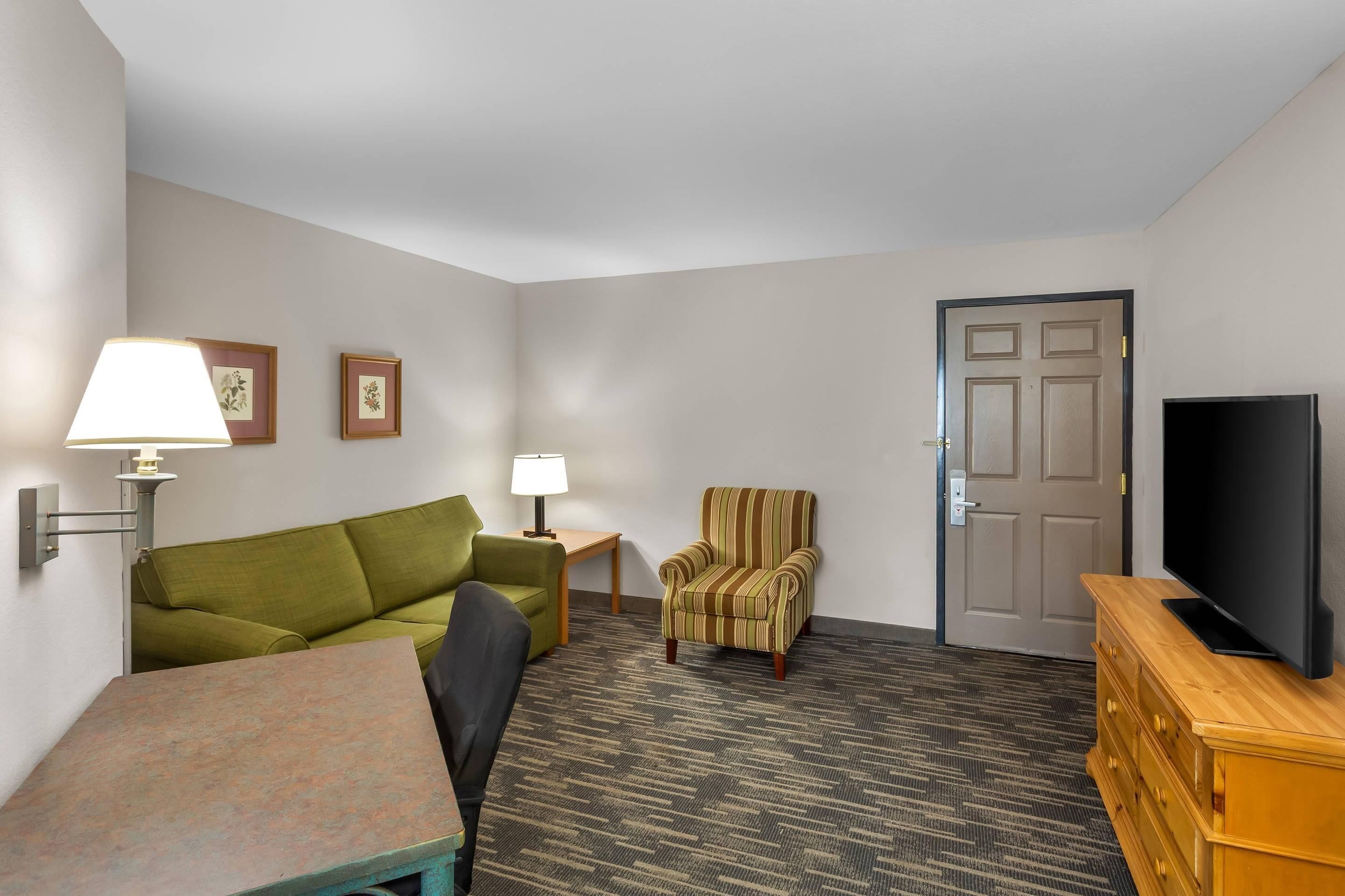 A hotel room with a green sofa, a striped armchair, a wooden TV stand, and a small table with a lamp.