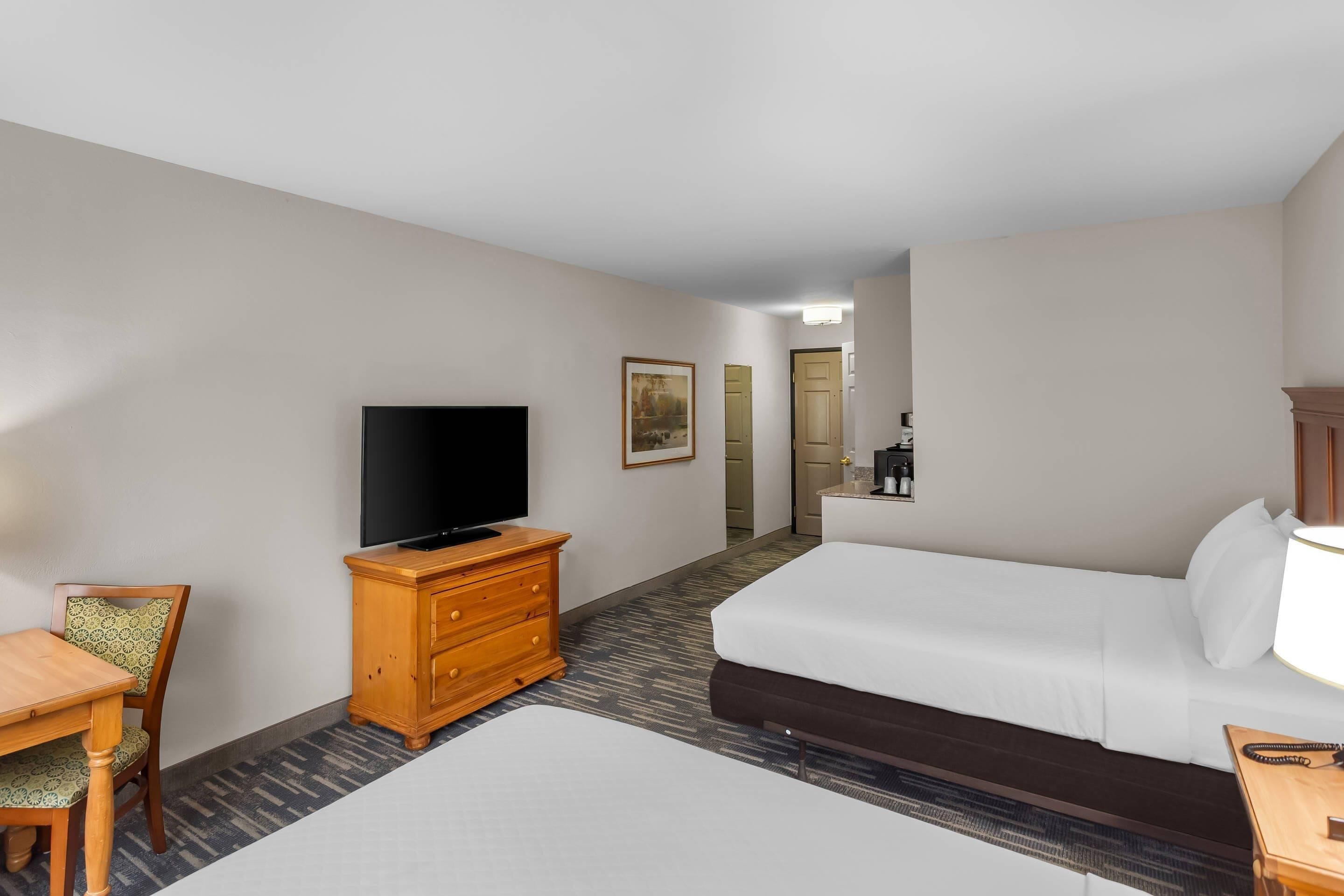 A hotel room with a bed, a television on a wooden dresser, a dining table with chairs, and a carpeted floor.