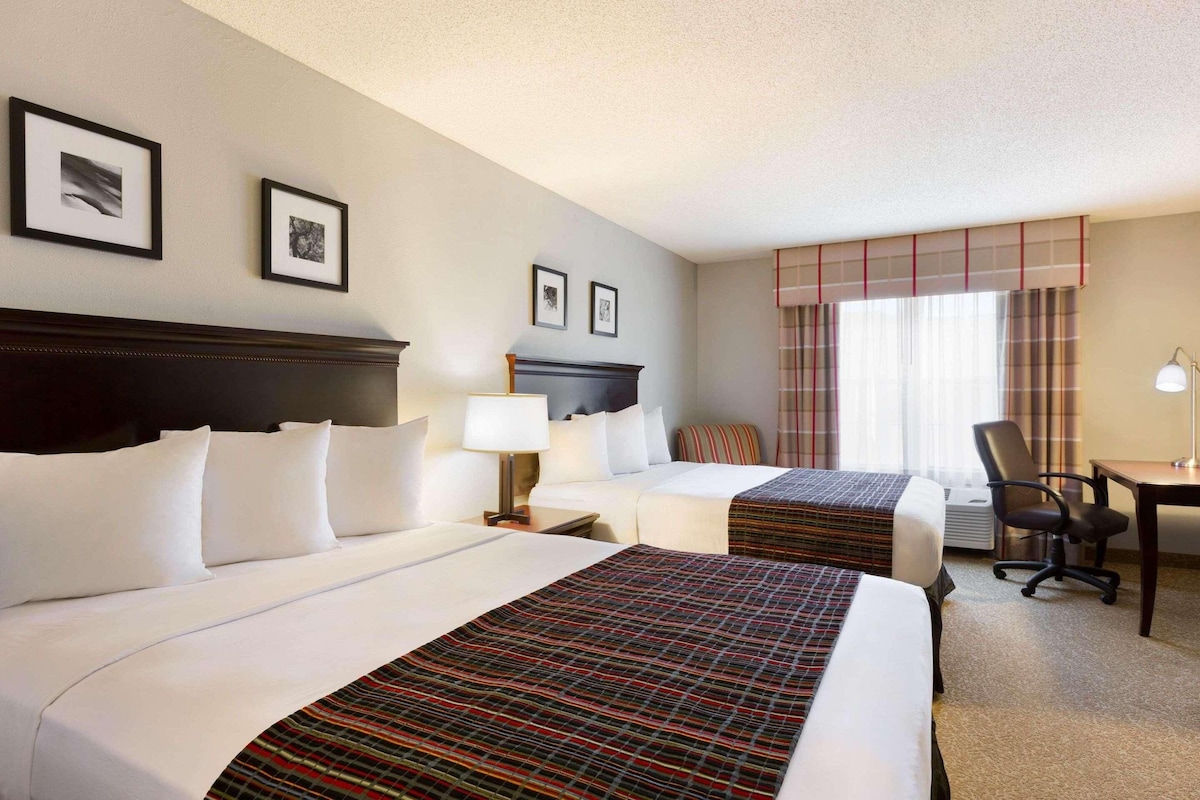 A hotel room with two beds, a desk, a chair, a lamp, and framed pictures on the wall.