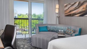 Habitación de hotel con una cama grande, un sofá, un escritorio y vistas a un balcón con vegetación.