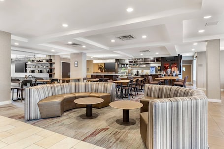 A modern hotel lobby with a seating area, a bar, and a kitchen in the background.