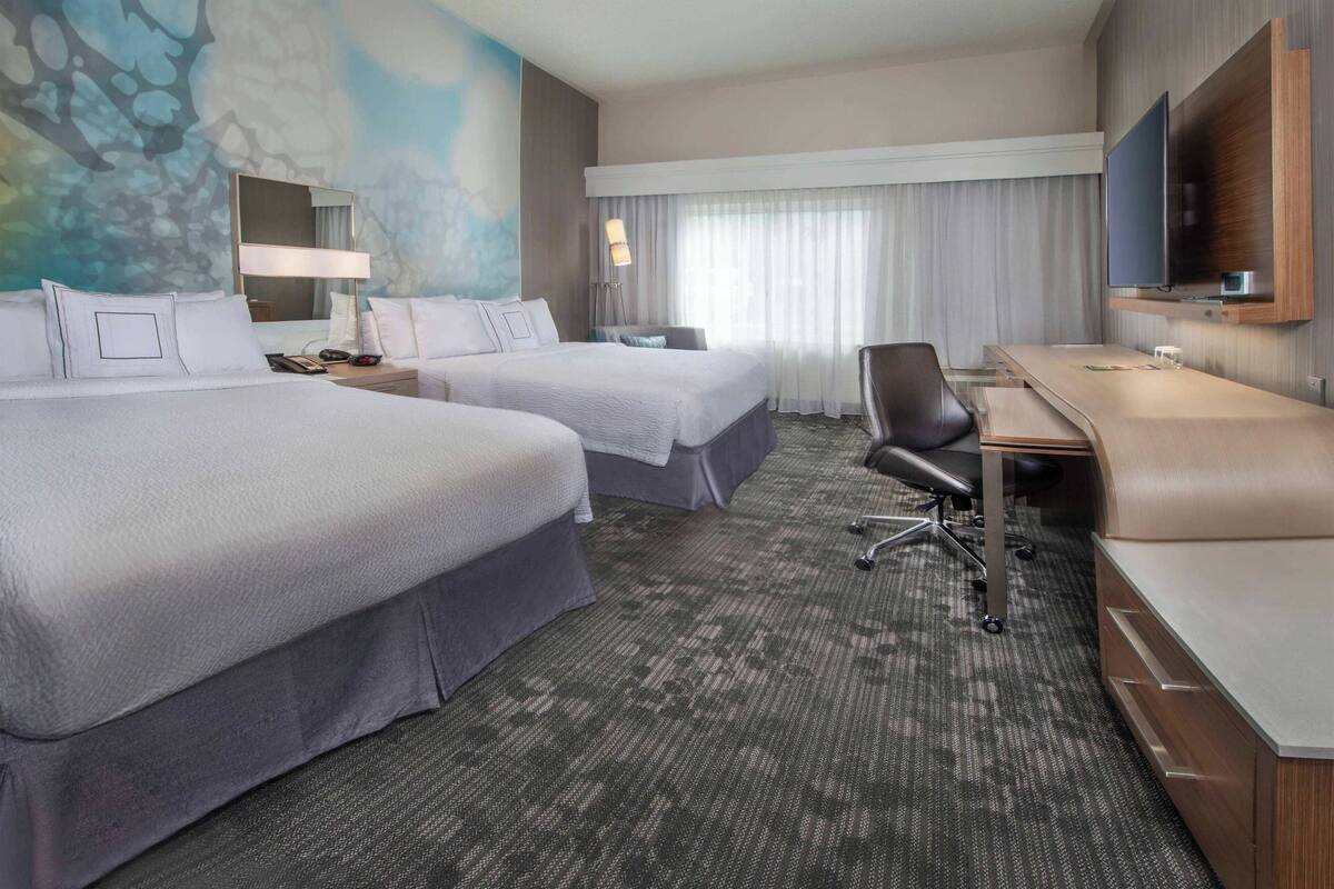A hotel room with two beds, a desk, a chair, a TV, and a large window with curtains.