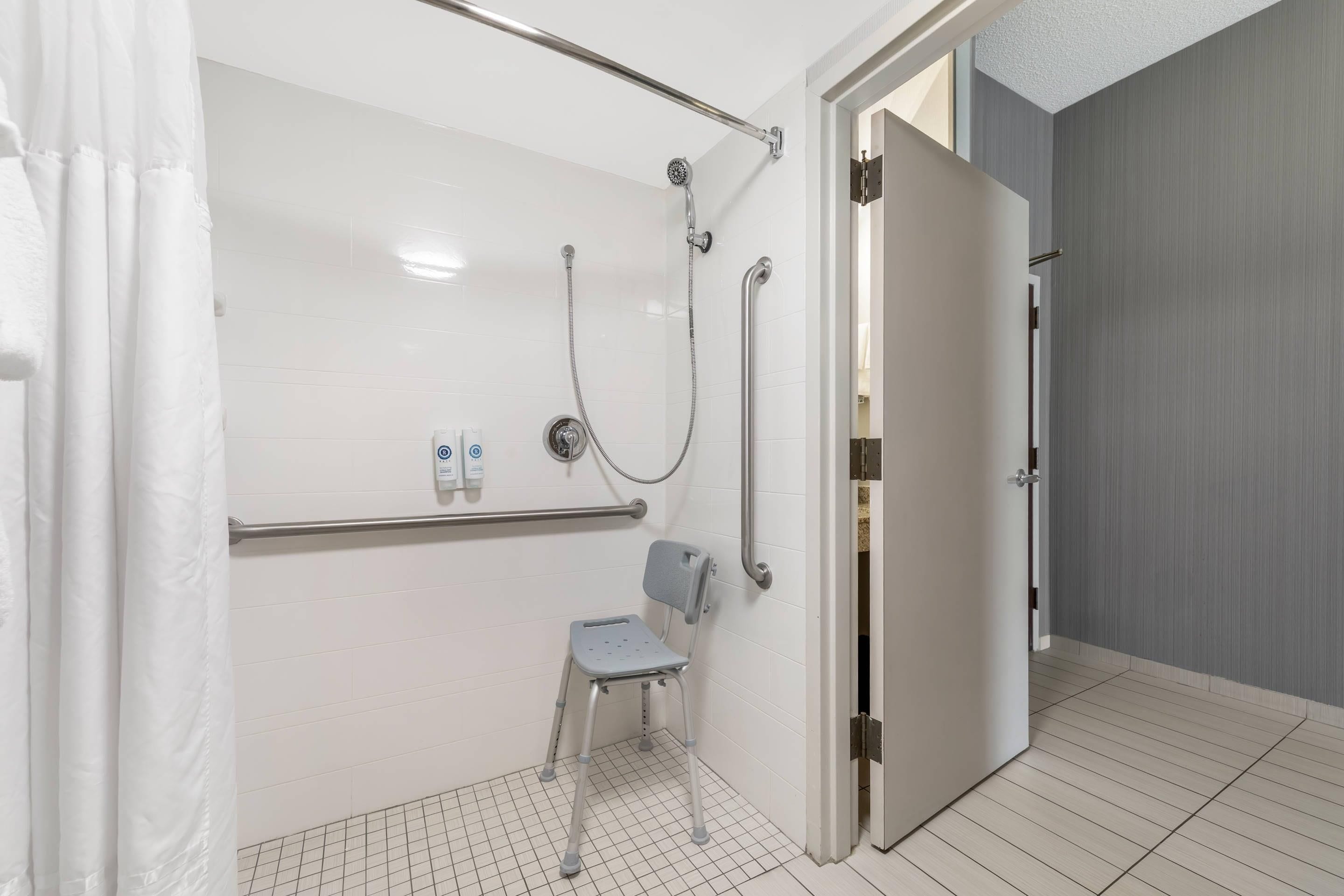 A modern bathroom with a shower, a grab bar, a wheelchair-accessible chair, and a door leading to another room.