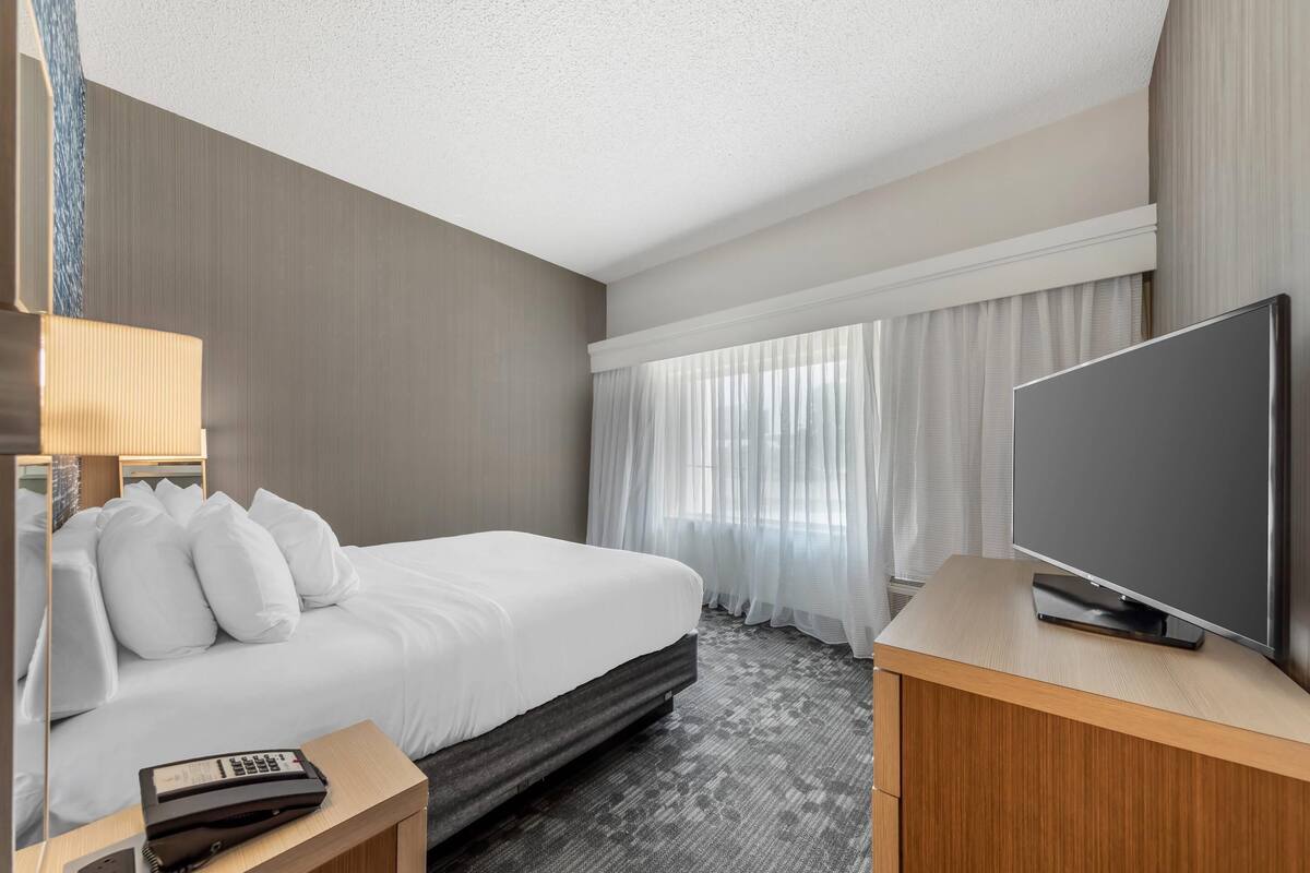 A hotel room with a bed, a TV, a bedside table, and a window with curtains.