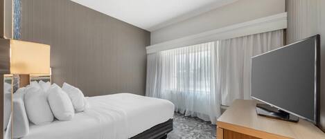Una habitación de hotel con cama, televisor, mesita de noche y ventana con cortinas.