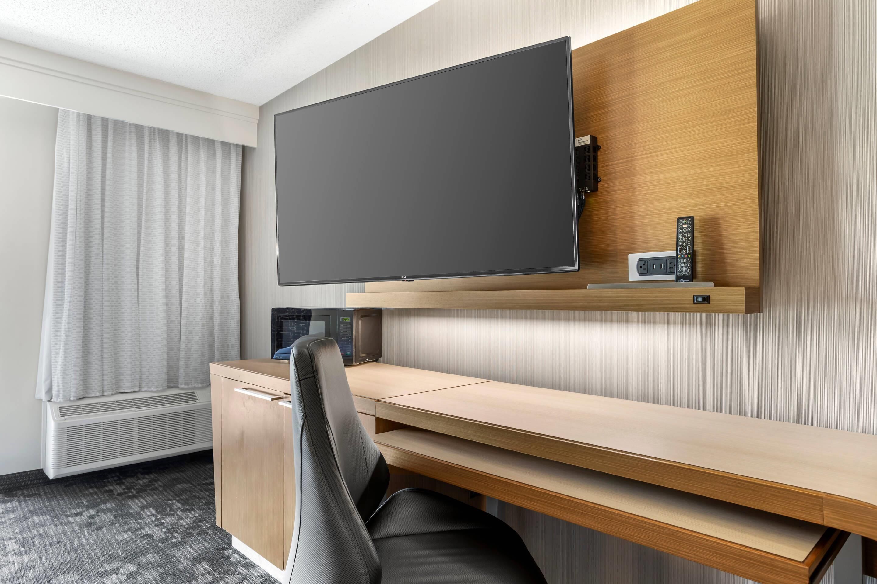A hotel room with a flat-screen TV mounted on a wooden wall, a desk with a chair, and a window with curtains.