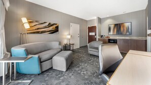 Una habitación de hotel moderna con un sofá gris, un sillón azul, una mesa de comedor de madera, un televisor de pantalla plana y una lámpara.