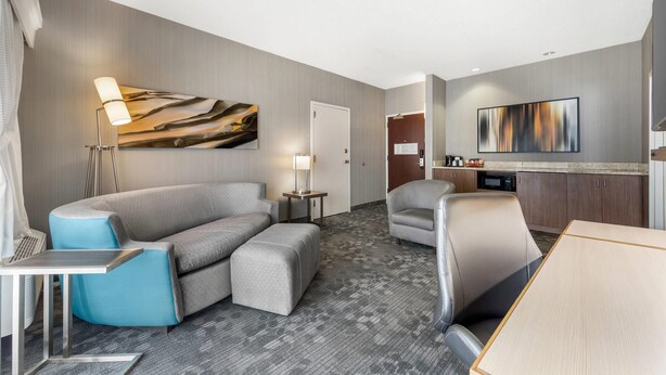 A modern hotel room with a grey sofa, a blue armchair, a wooden dining table, a flat-screen TV, and a lamp. - Comfort Inn & Suites Bethlehem Lehigh Valley Airport (Bethlehem)