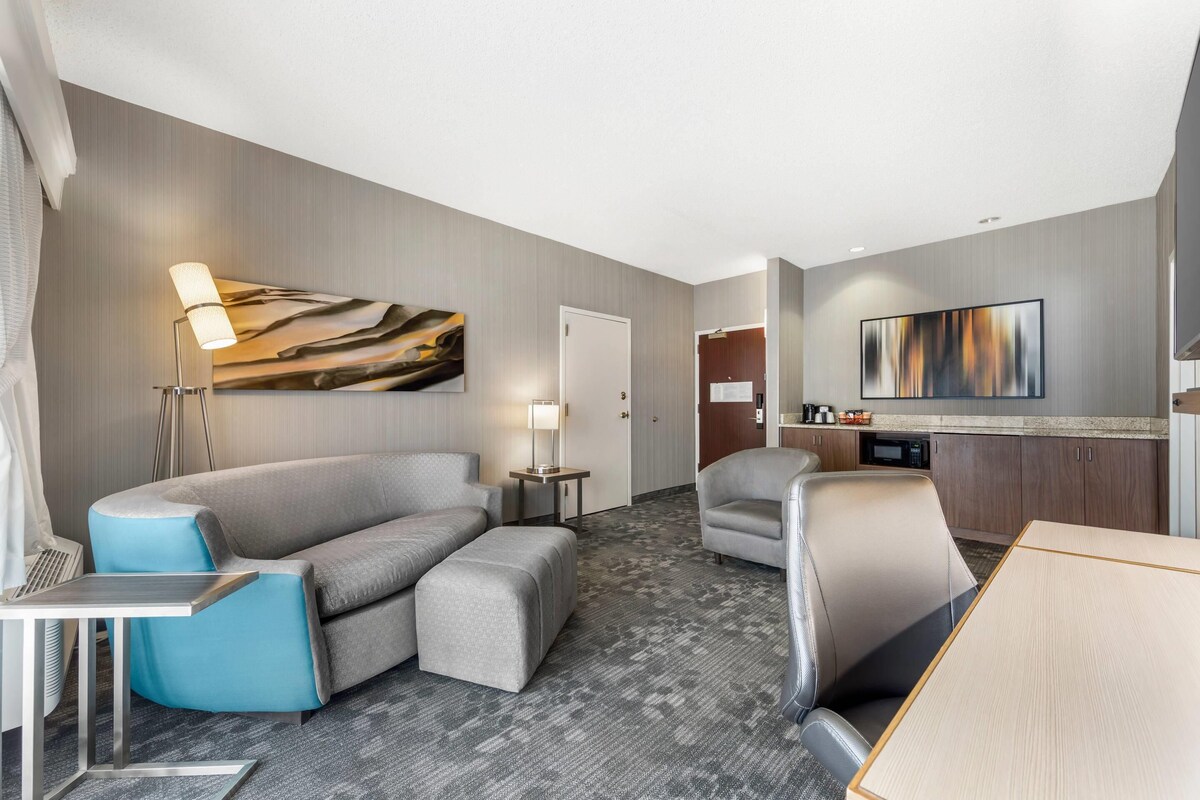 Una habitación de hotel moderna con un sofá gris, un sillón azul, una mesa de comedor de madera, un televisor de pantalla plana y una lámpara.