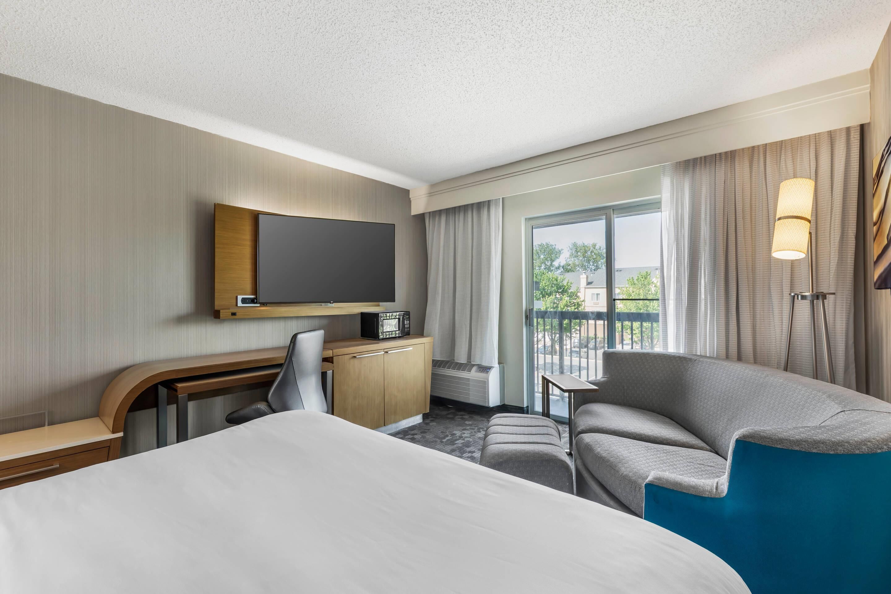 Habitación de hotel con cama, escritorio con televisor, sofá, sillón y balcón con cortinas.