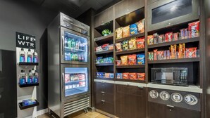 Un espacio de autoservicio con dispensadores refrigerados para bebidas, estantes con snacks, un microondas y una balanza.