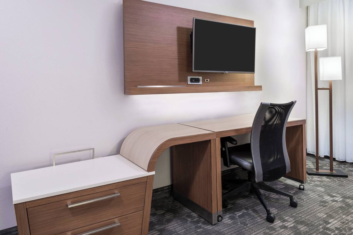 A modern hotel room with a wooden desk, a flat-screen TV mounted above it, a black office chair, and a lamp beside the desk.