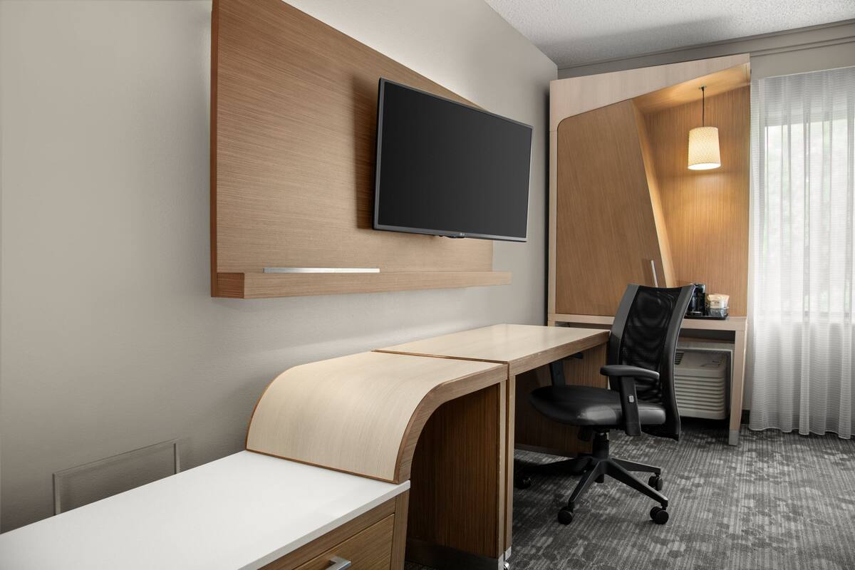 A modern hotel room with a flat-screen TV mounted on the wall, a curved desk, a black office chair, and a small desk with a lamp.