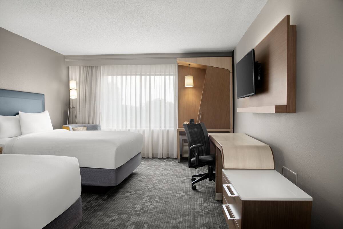 A modern hotel room with a large bed, a desk, a chair, a flat-screen TV mounted on the wall, and a window with curtains.