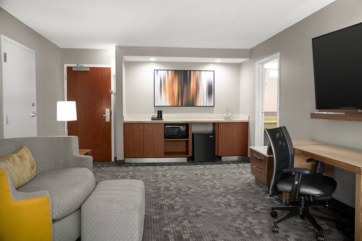 A compact hotel room with a kitchenette, a gray sofa with a yellow pillow, a black office chair, and a flat-screen TV mounted on the wall.