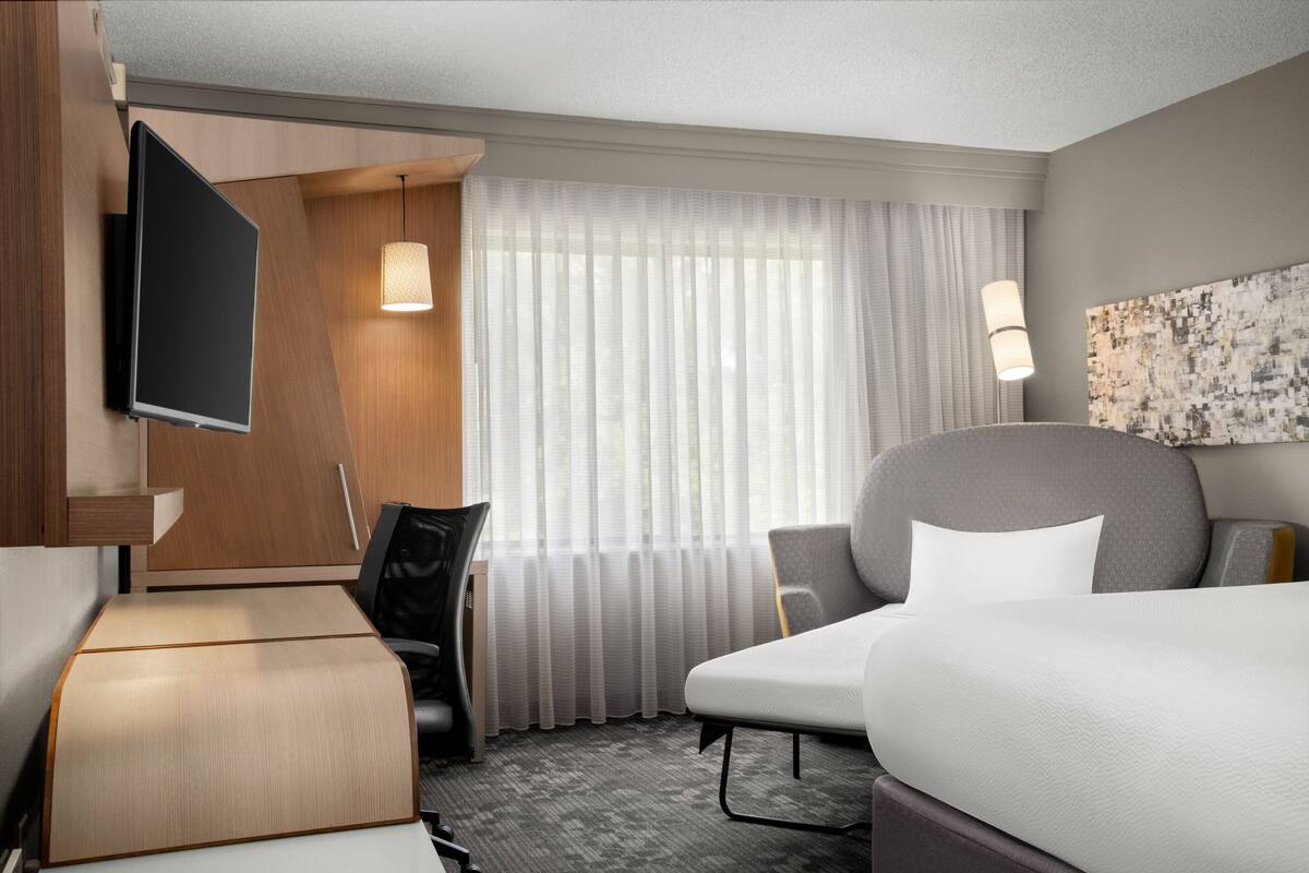 A hotel room with a bed, a desk, a chair, a TV, and a large window with curtains.