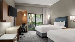 Quarto de hotel com duas camas, uma escrivaninha, uma cadeira, uma TV e uma porta de vidro deslizante para uma varanda.