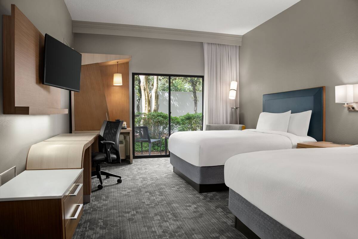 A hotel room with two beds, a desk, a chair, a TV, and a sliding glass door to a balcony.