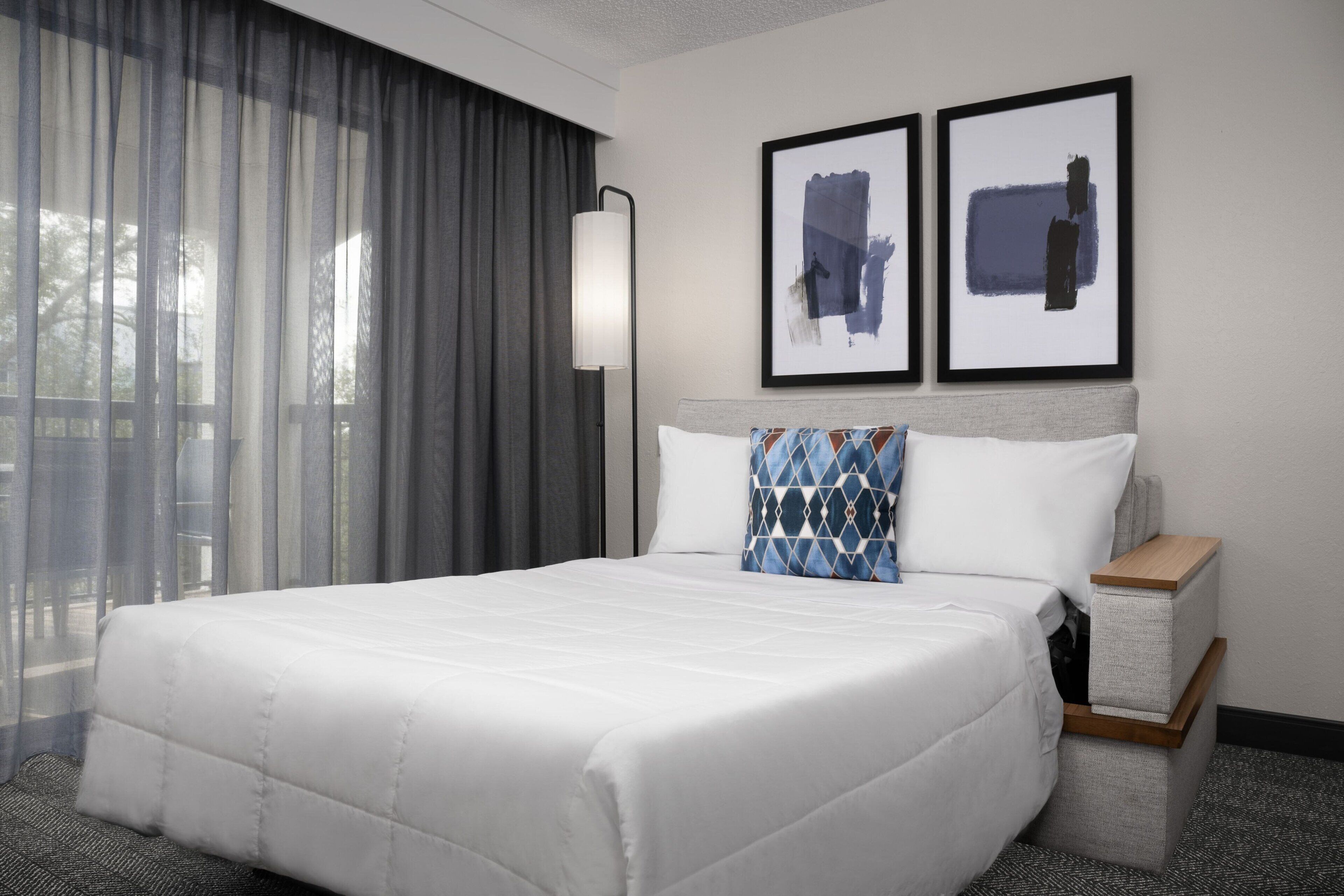 A hotel room with a large bed, a bedside table, a lamp, and two framed artworks on the wall.