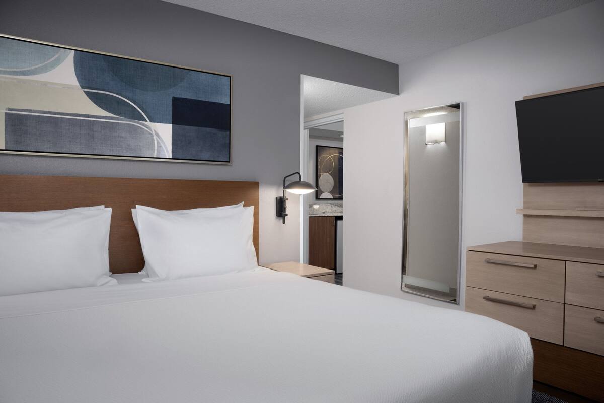 A hotel room with a large bed, a nightstand, a desk, a mirror, and a wall-mounted TV.