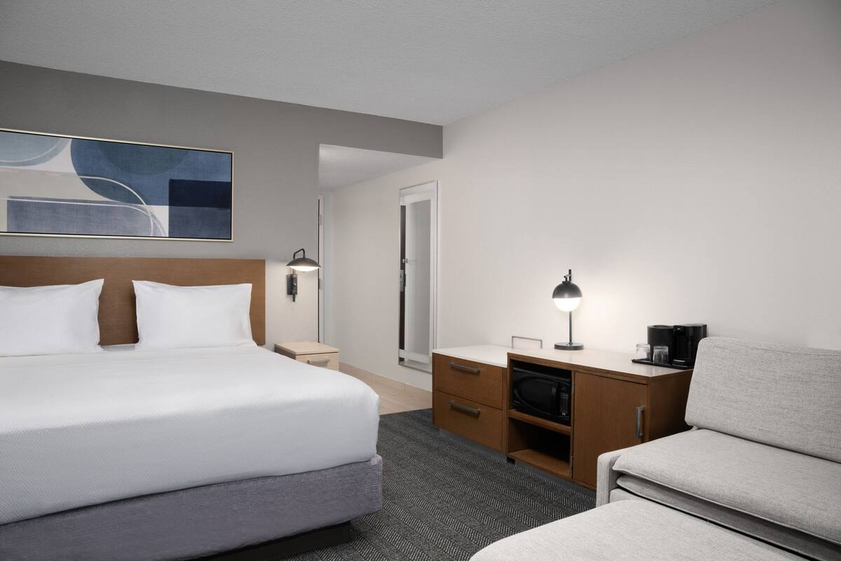 A hotel room with a large bed, a desk with a lamp, a chair, a wardrobe, and a mirror.