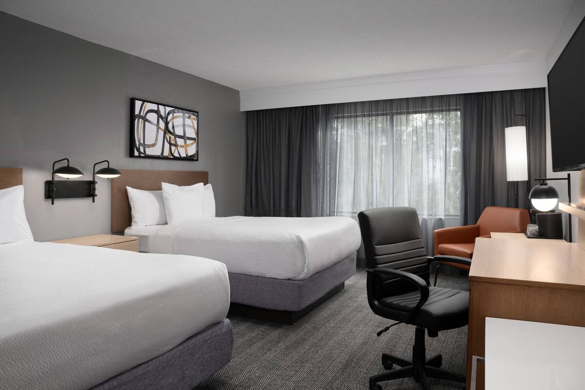 A hotel room with two beds, a desk, a chair, a TV, and a window with curtains.