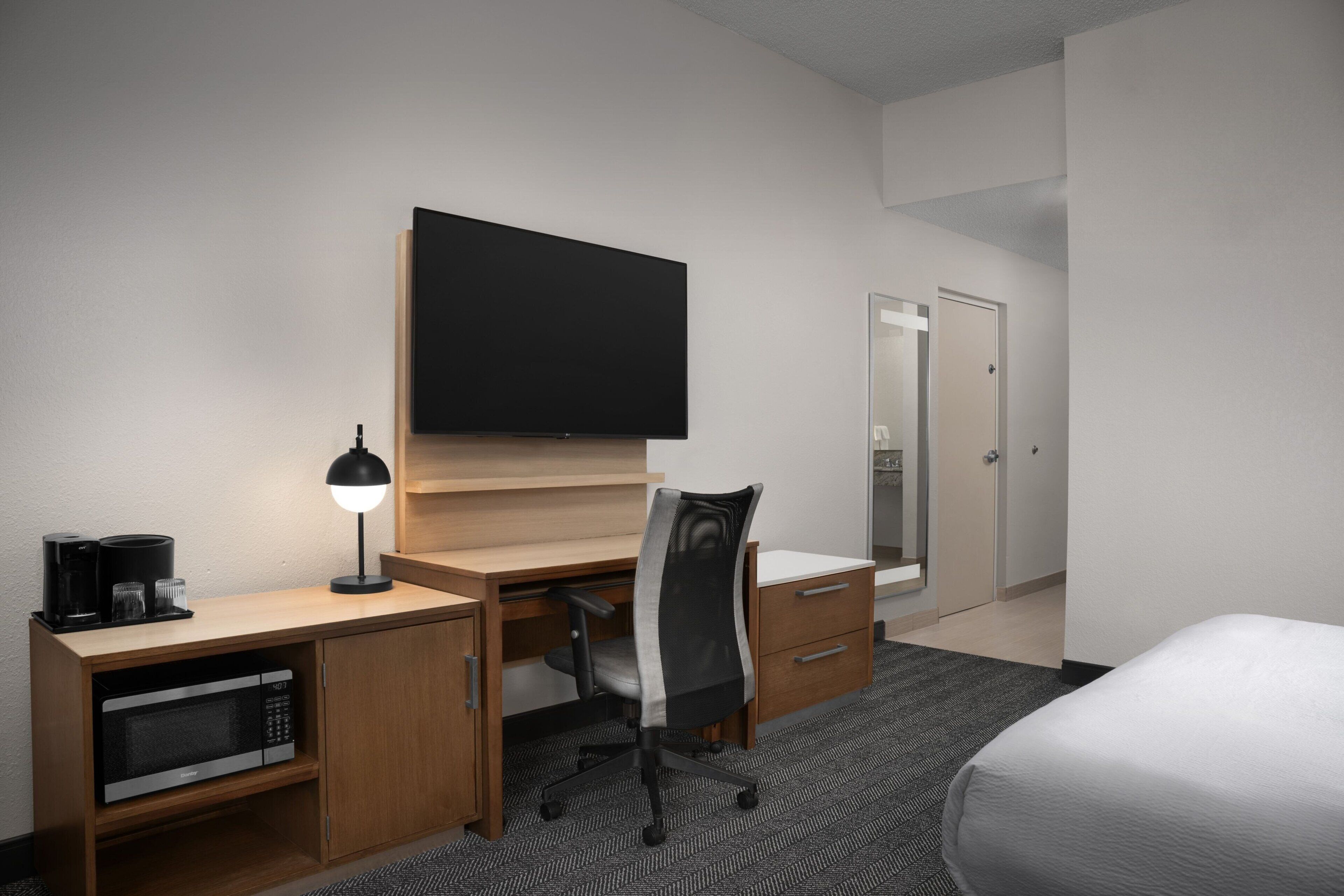 A hotel room with a bed, a desk with a computer, a chair, a flat-screen TV, a coffee maker, and a microwave.