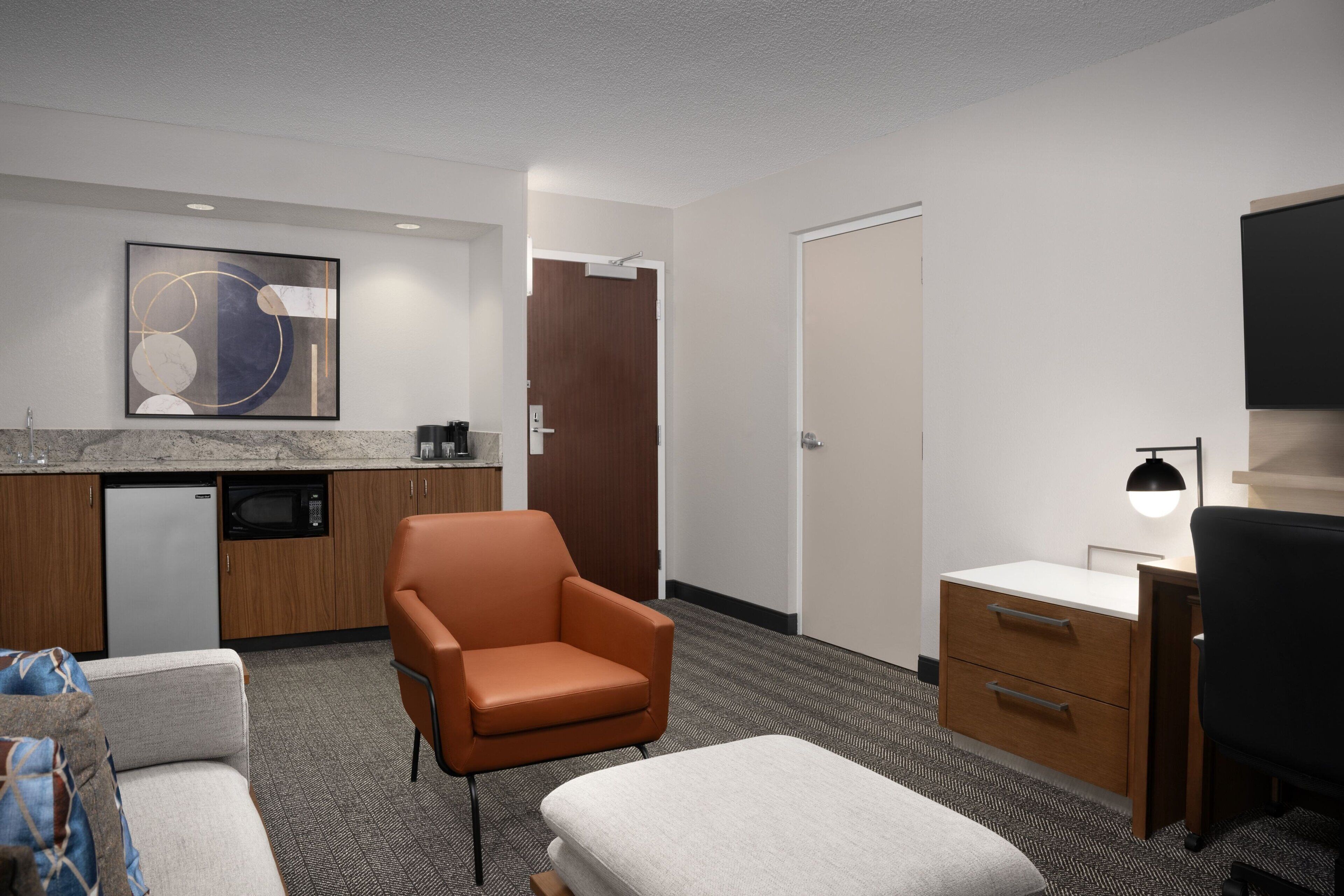 Quarto de hotel com cozinha compacta, sofá, poltrona, escrivaninha com luminária e televisão.