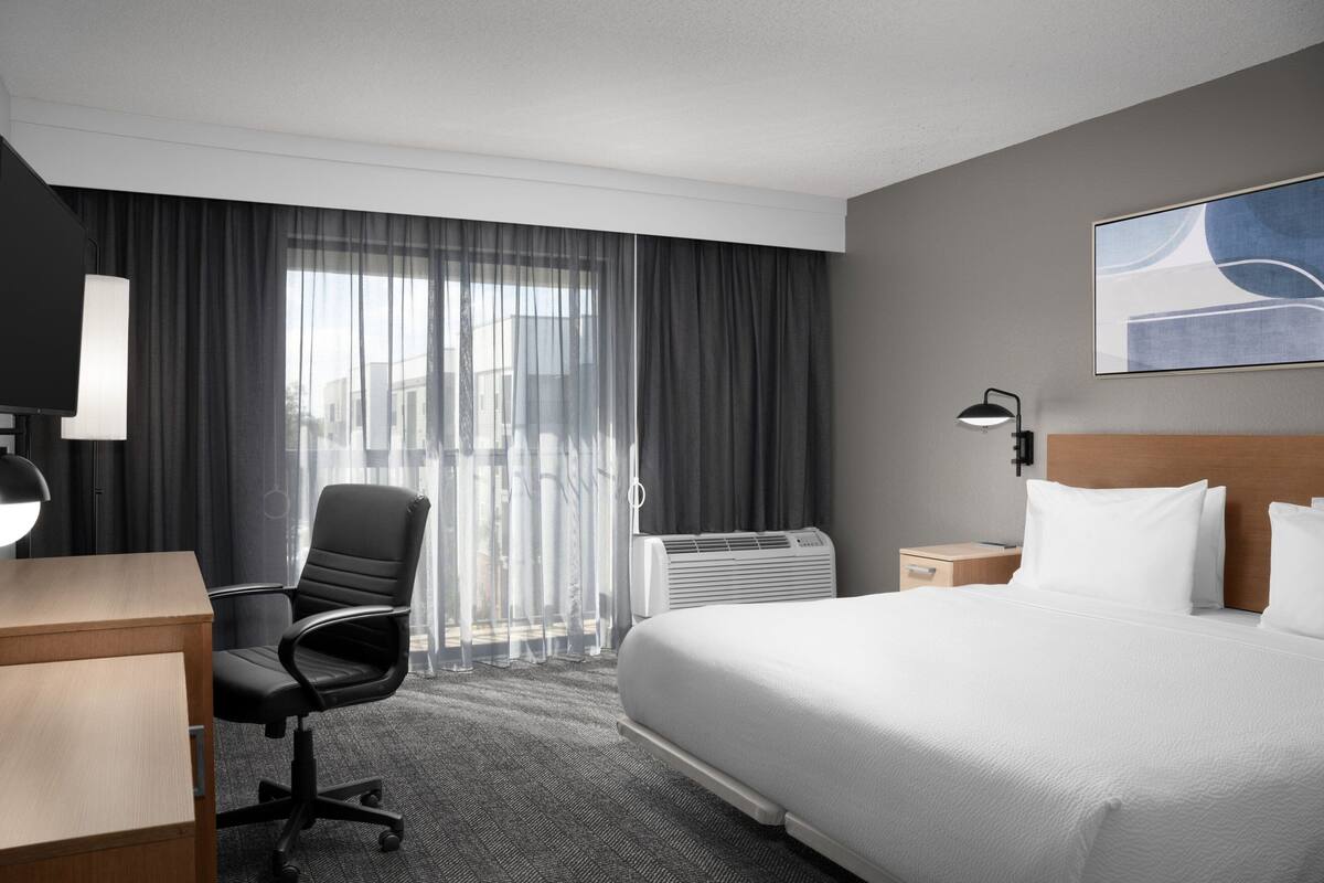 A hotel room with a bed, a desk, a chair, a window with curtains, an air conditioner, and a wall-mounted lamp.