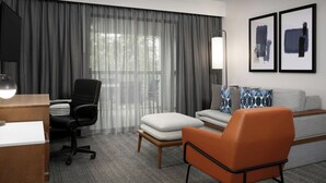 Una habitación de hotel con escritorio, silla de oficina, sofá, reposapiés y un ventanal con cortinas translúcidas.
