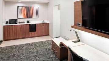 A modern hotel room with a kitchenette, a large flat-screen TV, a desk with a chair, and a carpeted floor.