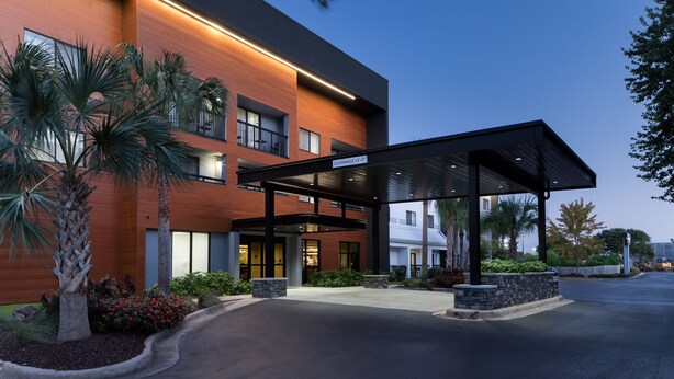 Una entrada de hotel moderna con una galería techada, palmeras y cielo despejado.