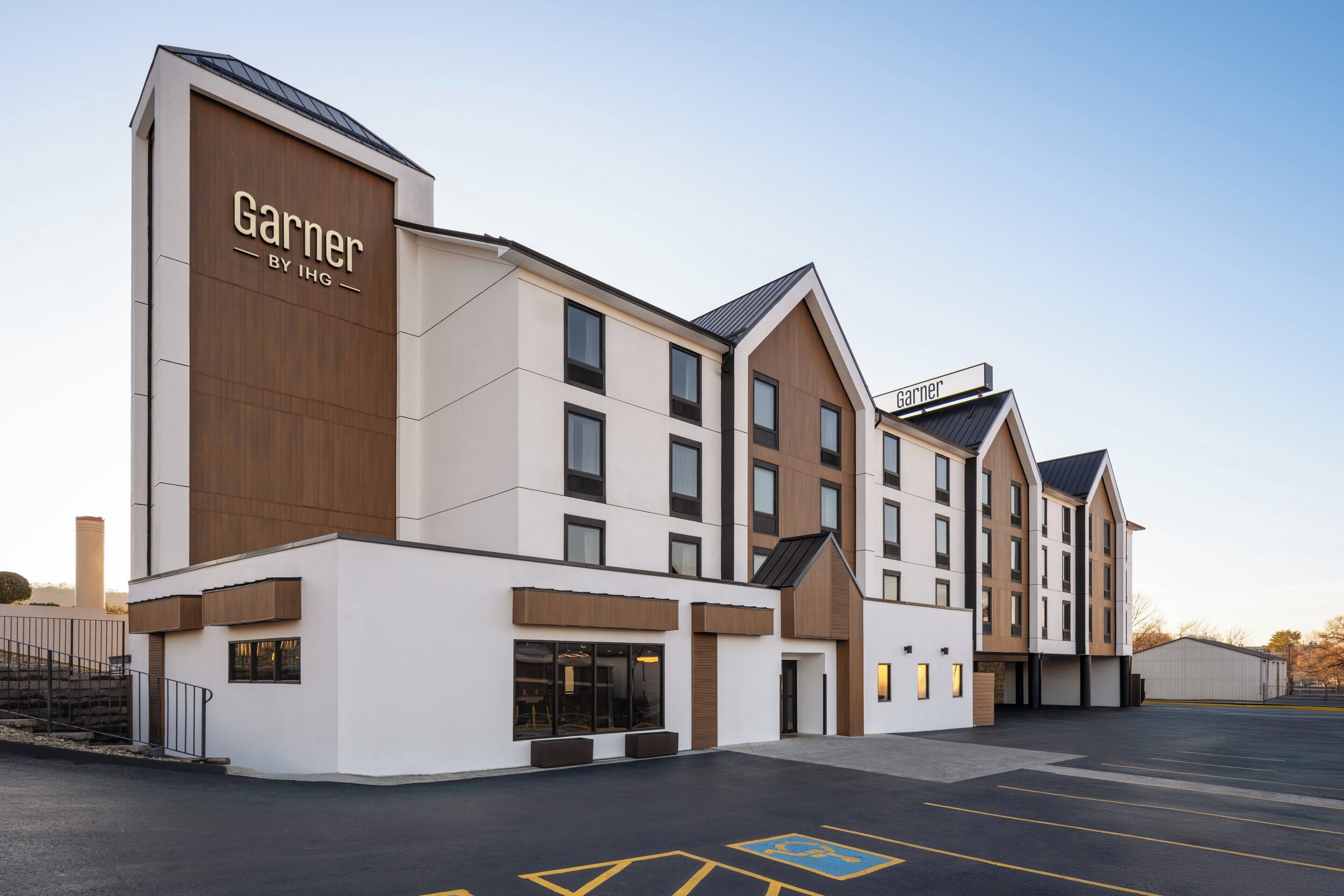 Un hôtel moderne arborant une enseigne bien visible « Garner by IHG », avec un mélange de panneaux blancs et en bois, de nombreux étages et un parking avec des places délimitées.