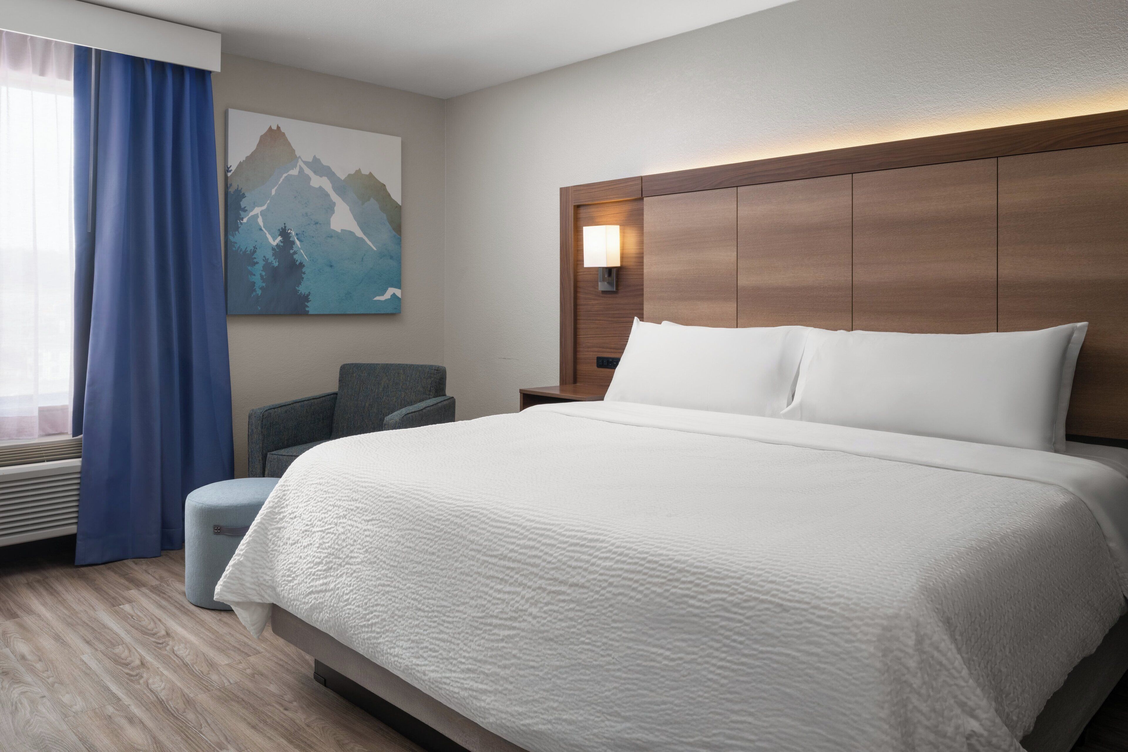 Une chambre d’hôtel avec un grand lit, une chaise, un tableau représentant des montagnes et une table de chevet avec une lampe.