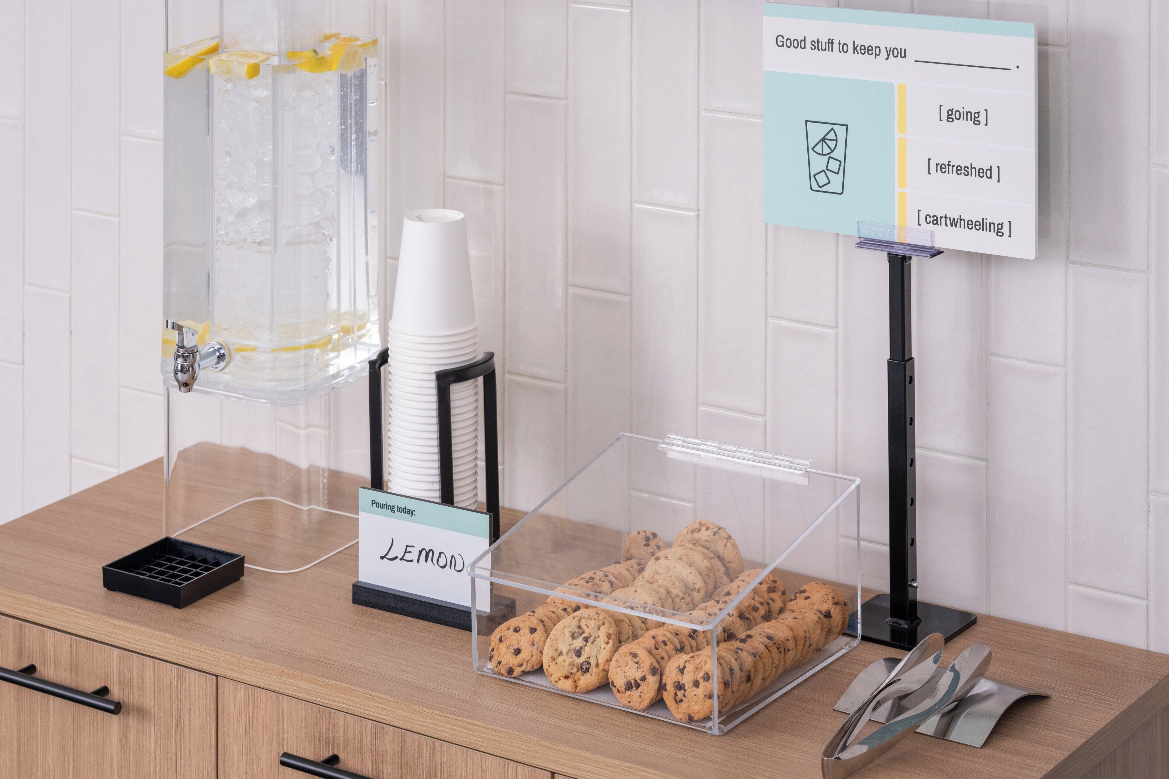 A self-serve station with lemon water, cookies, and a sign offering options like going, refreshed, and cartwheeling.