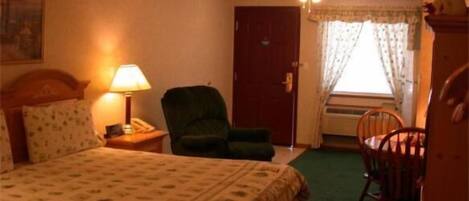 Quarto de hotel com uma cama, uma cadeira, uma escrivaninha com abajur e uma janela com cortinas.