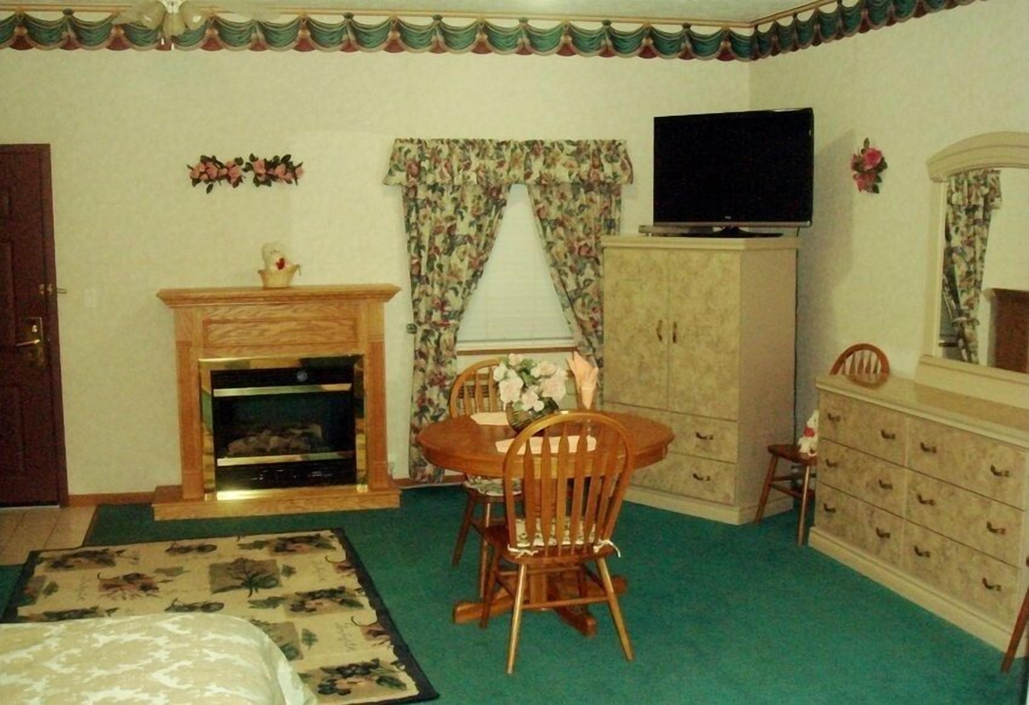 A bedroom with a bed, a wooden dresser, a television, a fireplace, and a dining table with chairs.